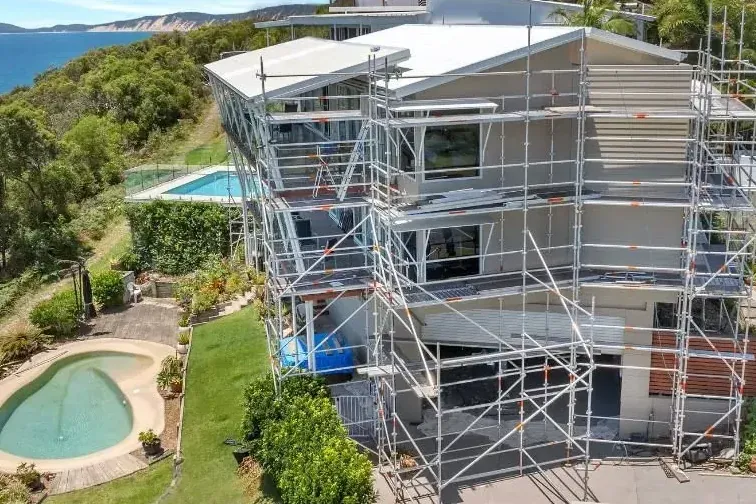 Aerial view of a multi-storey home under scaffolding on a coastal hillside, with the ocean behind