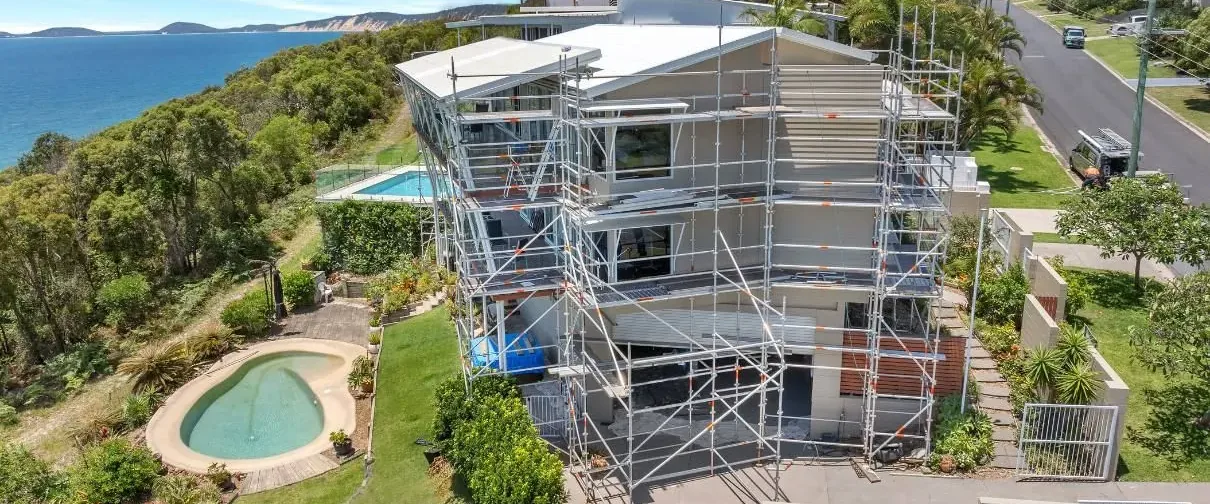 Aerial view of a multi-storey home under scaffolding on a coastal hillside, with the ocean behind